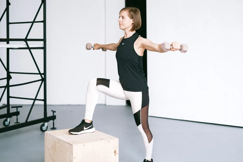 Woman holding dumbbells at shoulder height with one foot on a wooden exercise box